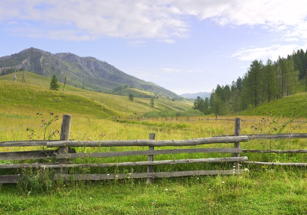 Ein Holzzaun auf einer grünen Wiese vor dem Hintergrund der Berge unter einem blau bewölkten Himmel Sibirien