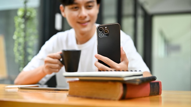 Ein hübscher asiatischer Mann, der sein Smartphone benutzt, während er einen Morgenkaffee an seinem Schreibtisch trinkt