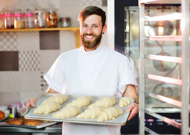 Ein hübscher Bäcker mit Bart bereitet Croissants zum Backen zu und lächelt in der Bäckerei