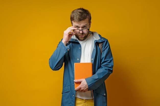 Ein hübscher bärtiger Student oder junger Wissenschaftler in einem Regenmantel und mit einem Buch hält eine Brille in der Hand