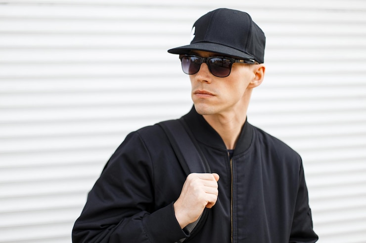 Foto ein hübscher, cooler, modischer hipster-mann mit stilvollen sonnenbrillen, baseballmütze, trendiger schwarzer jacke mit weißem, metallischem hintergrund, mann in schwarz.