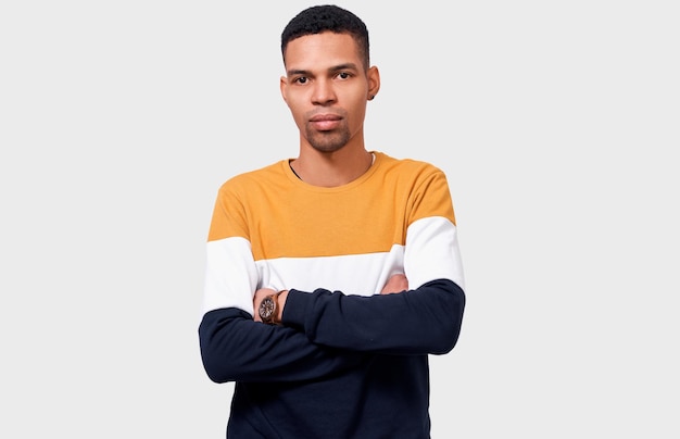 Ein hübscher, ernsthafter junger afroamerikanischer Mann, der einen trendigen Pullover trägt, hält die gekreuzten Hände, die über weißem Studiohintergrund auf die Kamera schauen Nachdenklicher männlicher Student, der an den Abschluss denkt Menschen