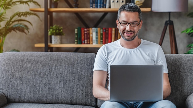 Ein hübscher, erwachsener Mann benutzt seinen Laptop, während er sich zu Hause auf dem Sofa entspannt