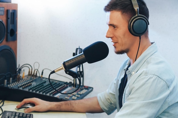 Ein hübscher, fröhlicher junger Radiomoderator mit Kopfhörern vor Mikrofon und Monitor nimmt im Studio eine Radiosendung auf. Daneben steht ein Mischpult und eine Tonsäule