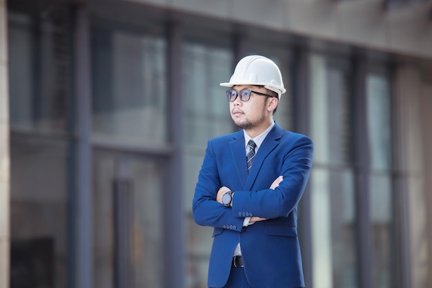 Ein hübscher Ingenieur in Anzug und Hut oder weißer Helm im Gebäudehintergrund, ein Ingenieur, der ein Tablet verwendet, um die Konstruktion eines modernen Gebäudes zu überprüfen.