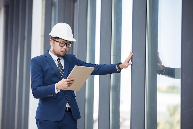 Ein hübscher Ingenieur in Anzug und Hut oder weißer Helm im Gebäudehintergrund, ein Ingenieur, der ein Tablet verwendet, um die Konstruktion eines modernen Gebäudes zu überprüfen.