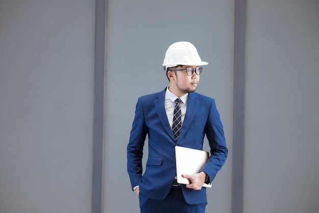 Ein hübscher Ingenieur in Anzug und Hut oder weißer Helm im Gebäudehintergrund, ein Ingenieur, der ein Tablet verwendet, um die Konstruktion eines modernen Gebäudes zu überprüfen.