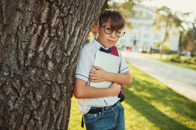 Ein hübscher Junge im schulpflichtigen Alter steht an einem Baum und hält eine Tafel. Der Schüler trägt Jeans und ein Hemd ohne Fliege.