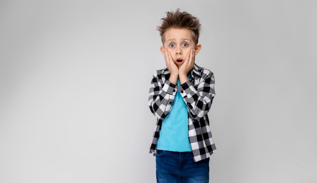 Ein hübscher Junge in einem karierten Hemd, in einem blauen Hemd und in Jeans steht auf Grau