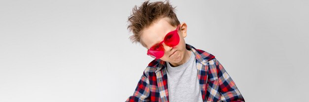 Ein hübscher Junge in einem karierten Hemd, in einem grauen Hemd und in Jeans steht auf einem grauen Hintergrund. Ein Junge mit roter Sonnenbrille. Der Junge zieht seine Hände nach vorne.