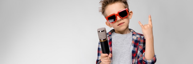 Ein hübscher Junge in einem karierten Hemd, in einem grauen Hemd und in Jeans steht auf einer grauen Wand.