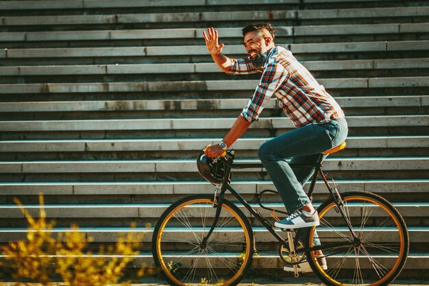 Ein hübscher junger Mann fährt mit seinem Fahrrad in die Stadt, setzt sich darauf und winkt jemandem zu.