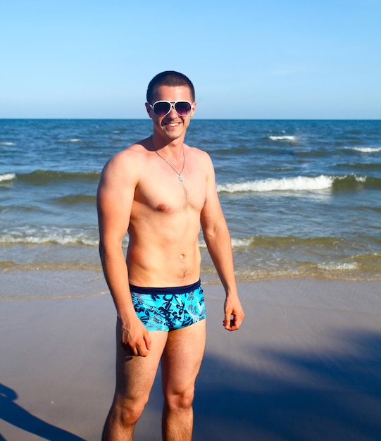 Foto ein hübscher junger mann in badehose und sonnenbrille, der im sand steht, mit ozean im hintergrund