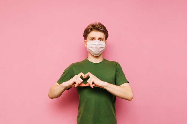 Ein hübscher junger Mann in einer medizinischen Maske steht auf einem rosafarbenen Hintergrund, schaut in die Kamera und zeigt eine Herzgeste Guy in einer schützenden Gazemaske zeigt eine Geste der LiebeCoronavirus-Pandemie