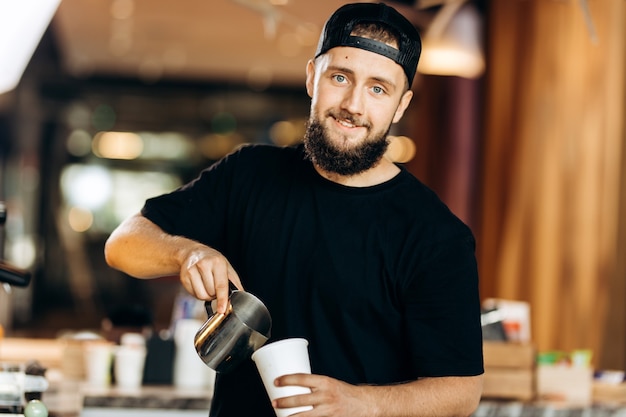 Ein hübscher junger Mann mit Bart, der in lässigem Stil gekleidet ist, fügt dem Kaffee in einem modernen Café Milch hinzu. .