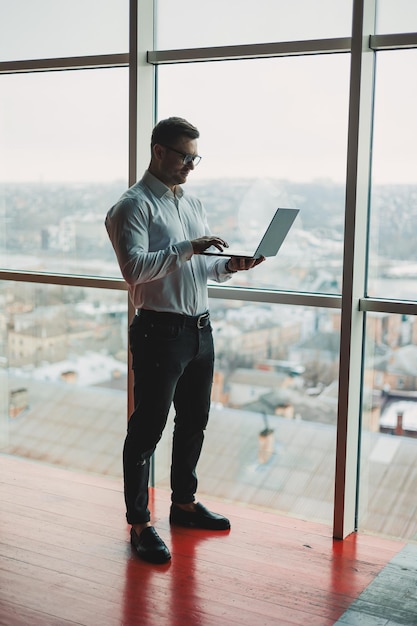 Ein hübscher junger Mann mit einem Laptop steht auf dem Hintergrund eines großen Panoramafensters im Himmel. Ein modernes Büro mit großen Fenstern und ein Büroangestellter