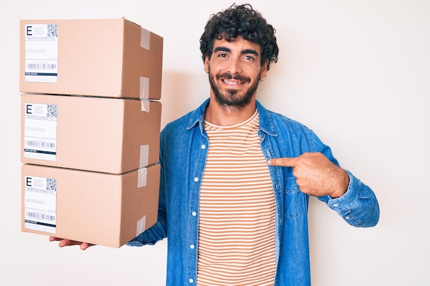 Ein hübscher junger Mann mit lockigem Haar und Bären hält ein Lieferpaket, zeigt mit dem Finger auf ein Selbst, lächelt glücklich und stolz