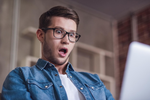 Ein hübscher Kerl in lässiger Kleidung und Brille benutzt einen Laptop und zeigt Überraschung, während er sitzt