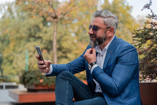 Ein hübscher, stilvoller, reifer Geschäftsmann, der einen Videoanruf auf seinem Telefon tätigt, während er auf einer Parkbank sitzt