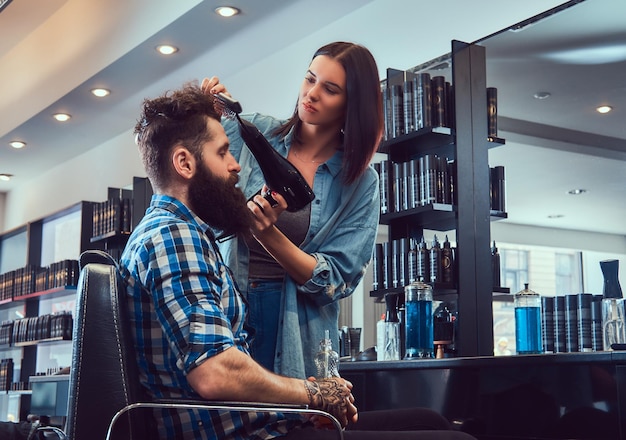 Ein hübscher, stylischer bärtiger Mann mit einem Tattoo auf dem Arm, gekleidet in ein Flanellhemd, das Saft hält, während die Friseurfrau einen Fön in einem Friseursalon benutzt.