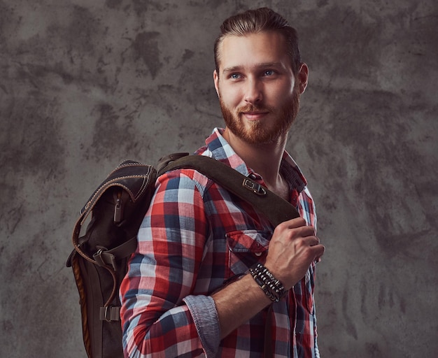 Ein hübscher, stylischer rothaariger Reisender in einem Flanellhemd mit Rucksack, der in einem Studio auf grauem Hintergrund posiert.