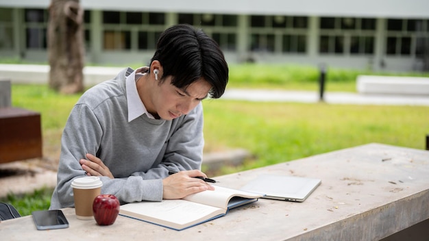 Ein hübscher und schlauer asiatischer College-Mann konzentriert sich auf das Lesen eines Buches im Campuspark