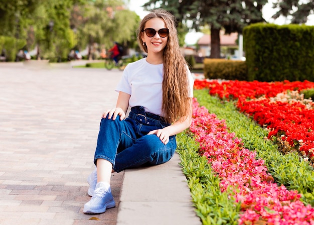 Ein hübsches junges Mädchen in einem weißen T-Shirt und Jeans mit Sonnenbrille sitzt in der Nähe eines Blumenbeets