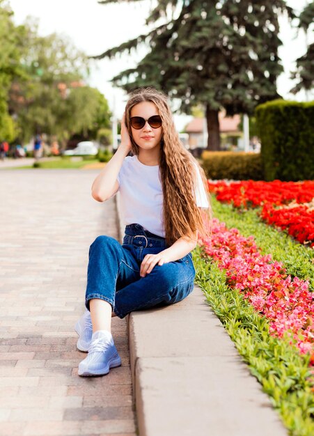 Ein hübsches junges Mädchen in einem weißen T-Shirt und Jeans mit Sonnenbrille sitzt in der Nähe eines Blumenbeets