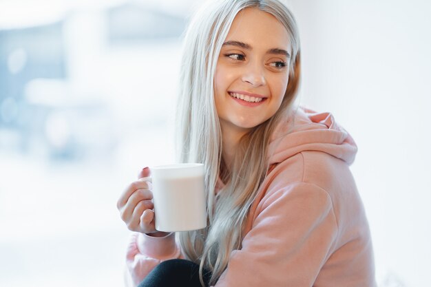 Ein hübsches lächelndes Mädchen, das eine Tasse Kaffee oder Milch genießt, während es durch das verschwommene Fensterglas sitzt. Trägt einen lässigen rosa, blassen Parka. Schließen Sie oben weiße Tasse in Frauenhänden nahe Lippen.