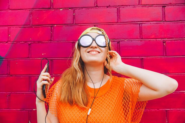 Ein hübsches Mädchen steht in der Nähe einer orangefarbenen Ziegelwand mit Kopfhörern, die ein Mobiltelefon halten. Ein Porträt eines jungen Mädchens im Teenageralter, das Musik hört