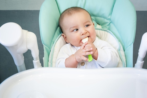 Ein hungriges Baby nagt an einem Plastiklöffel am Tisch auf einem Hochstuhl. Kinderkrankheiten. Juckendes Zahnfleisch. Einführung von Beikost