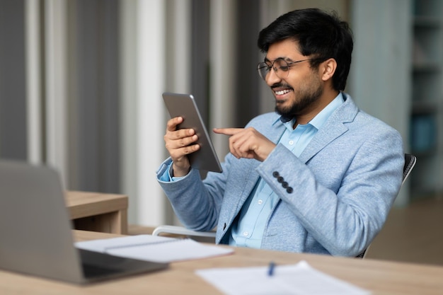 Ein indischer Geschäftsmann sitzt an einem modernen Büro-Schreibtisch und surft drinnen auf einem Tablet
