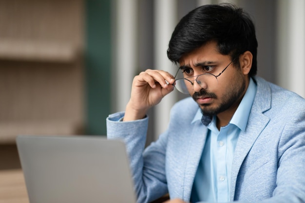 Ein indischer Mann, der mit Sehproblemen zu tun hat, zwickelt beim Laptop im Innenraum.