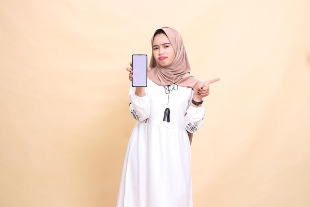 Ein indonesisches muslimisches Mädchen, das einen Hijab trägt, runzt die Stirn und zeigt einen Handy-Bildschirm, der nach links zeigt.