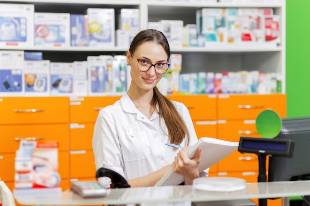 Ein jugendliches, angenehmes dunkelhaariges Mädchen mit Brille, gekleidet in einen medizinischen Overall, schreibt an der Kasse einer neuen Apotheke Notizen in ein Notizbuch. .