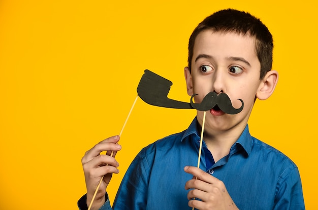 Ein Junge in einem blauen Hemd auf gelbem Grund verzieht das Gesicht und kleidet einen Schnurrbart und hält eine Pfeife