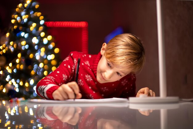 Ein Junge in einem roten Weihnachtspullover auf dem Hintergrund eines Weihnachtsbaums malt etwas in seinem Zimmer