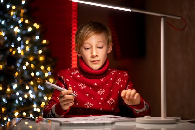 Ein Junge in einem roten Weihnachtspullover auf dem Hintergrund eines Weihnachtsbaums sitzt in seinem Kinderzimmer und zeichnet