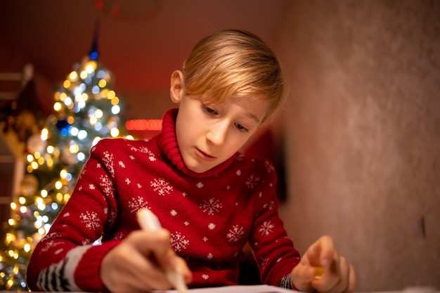 Ein Junge in einem roten Weihnachtspullover auf dem Hintergrund eines Weihnachtsbaums zeichnet in seinem Zimmer unter einer Lampe