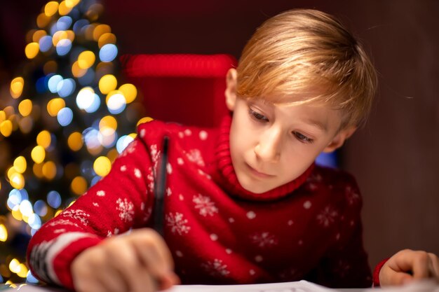 Ein Junge in einem roten Weihnachtspullover vor dem Hintergrund eines Weihnachtsbaums sitzt an einem Tisch in einem Raum