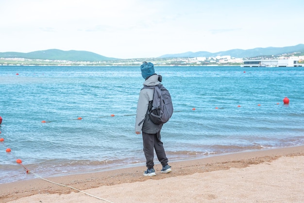 Ein Junge in einer grauen Jacke spielt mit Sand am Strand. Der Schuljunge berührt im Herbst das Wasser