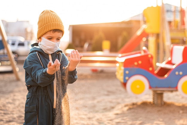 Ein Junge in einer Schutzmaske spielt auf dem Spielplatz und verschüttet während der Pandemie von Coronavirus und Covid - 19 Sand aus seinen Händen