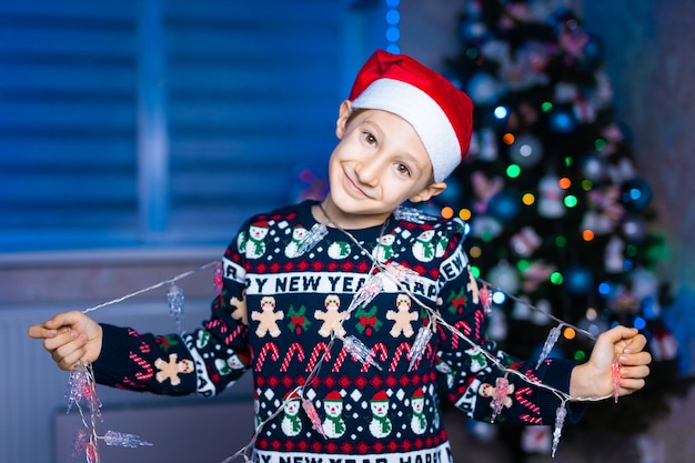 ein Junge in einer Weihnachtsmannmütze mit Girlanden in seinen Händen auf dem Hintergrund eines Weihnachtsbaumes mit Neonlicht