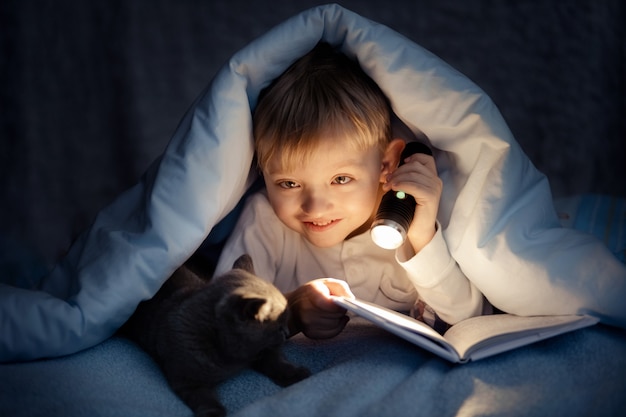 Ein Junge liest abends im Dunkeln unter einer Decke mit einer Katze ein Buch