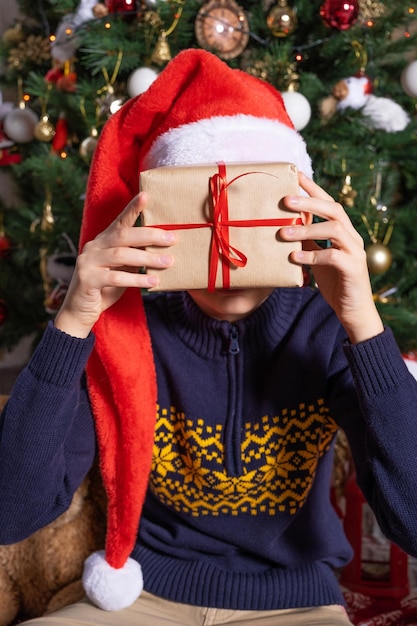 Ein Junge mit Weihnachtsmannmütze verbirgt sein Gesicht hinter einem verpackten Weihnachtsgeschenk