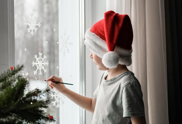 Ein Junge mit Weihnachtsmütze malt Schneeflocken ans Fenster. Familientraditionen zu Weihnachten