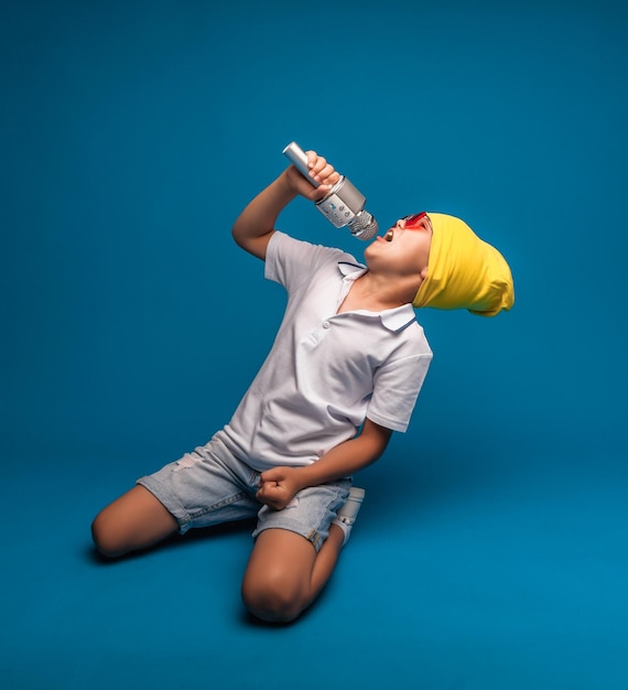 Ein Junge singt auf blauem Hintergrund in ein Mikrofon Ein hübscher Junge in weißem T-Shirt und Shorts steht auf blauem Hintergrund und singt emotional ins Mikrofon