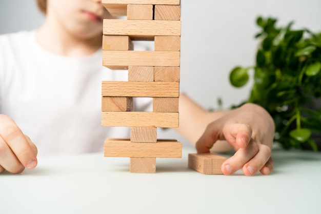 Ein Junge spielt ein Brettspiel und baut einen Turm aus Holzwürfeln. Hände schließen ein logisches Spiel von Jenga für