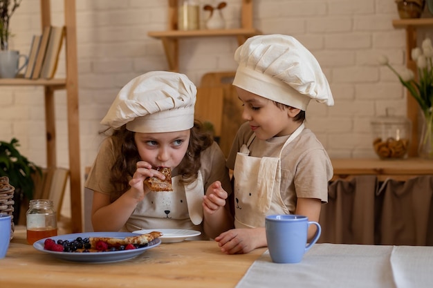 Ein Junge und ein blondes Mädchen mit Schürze und Kochmütze am Tisch essen Pfannkuchen