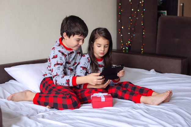 Ein Junge und ein Mädchen im roten und grauen Pyjama sitzen zu Hause auf dem Bett und lächeln auf ein digitales Tablet